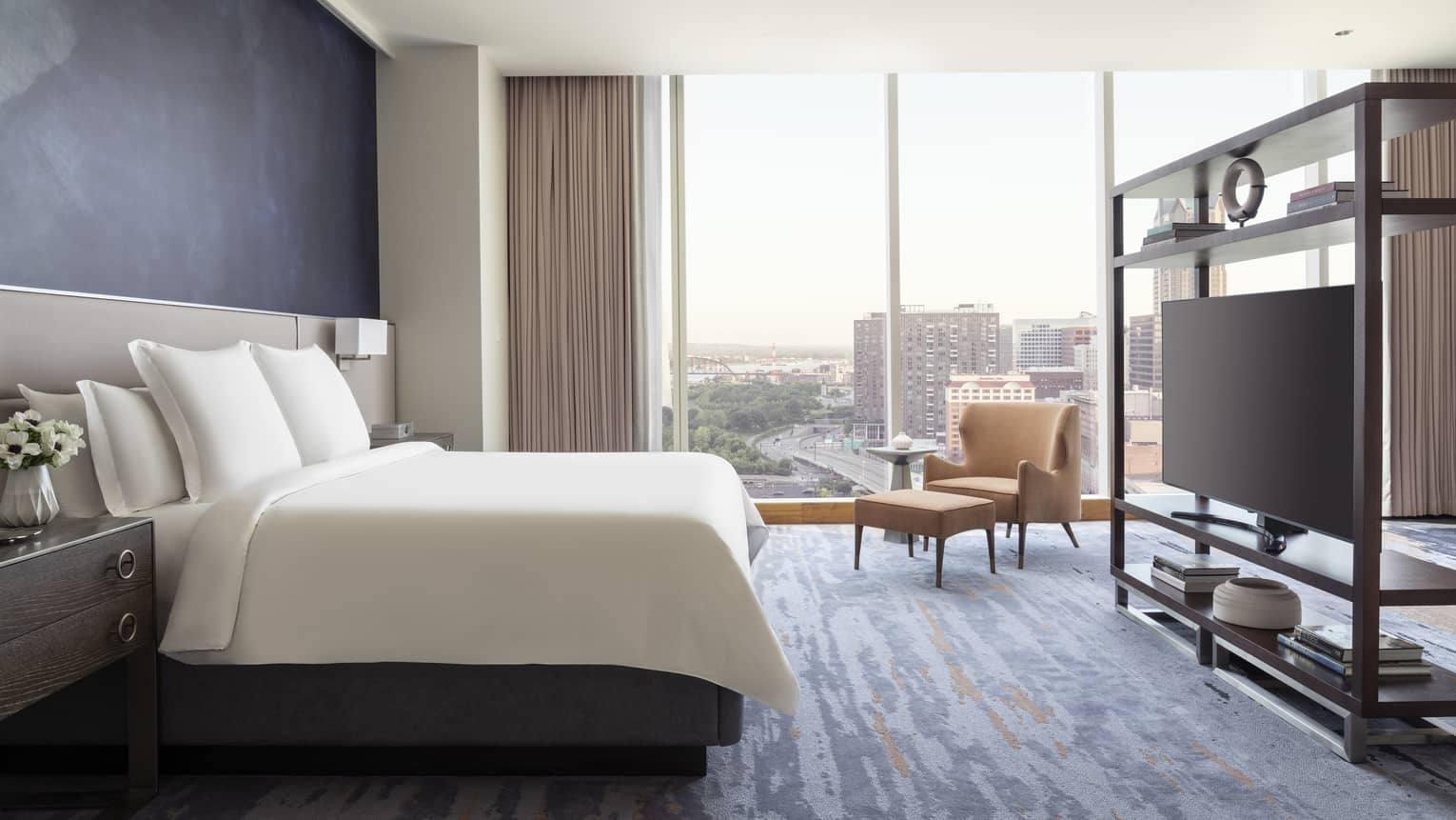Luxury hotel room with a king bed, armchair and ottoman, shelving unit with a flat-screen TV, and floor-to-ceiling windows with a city view