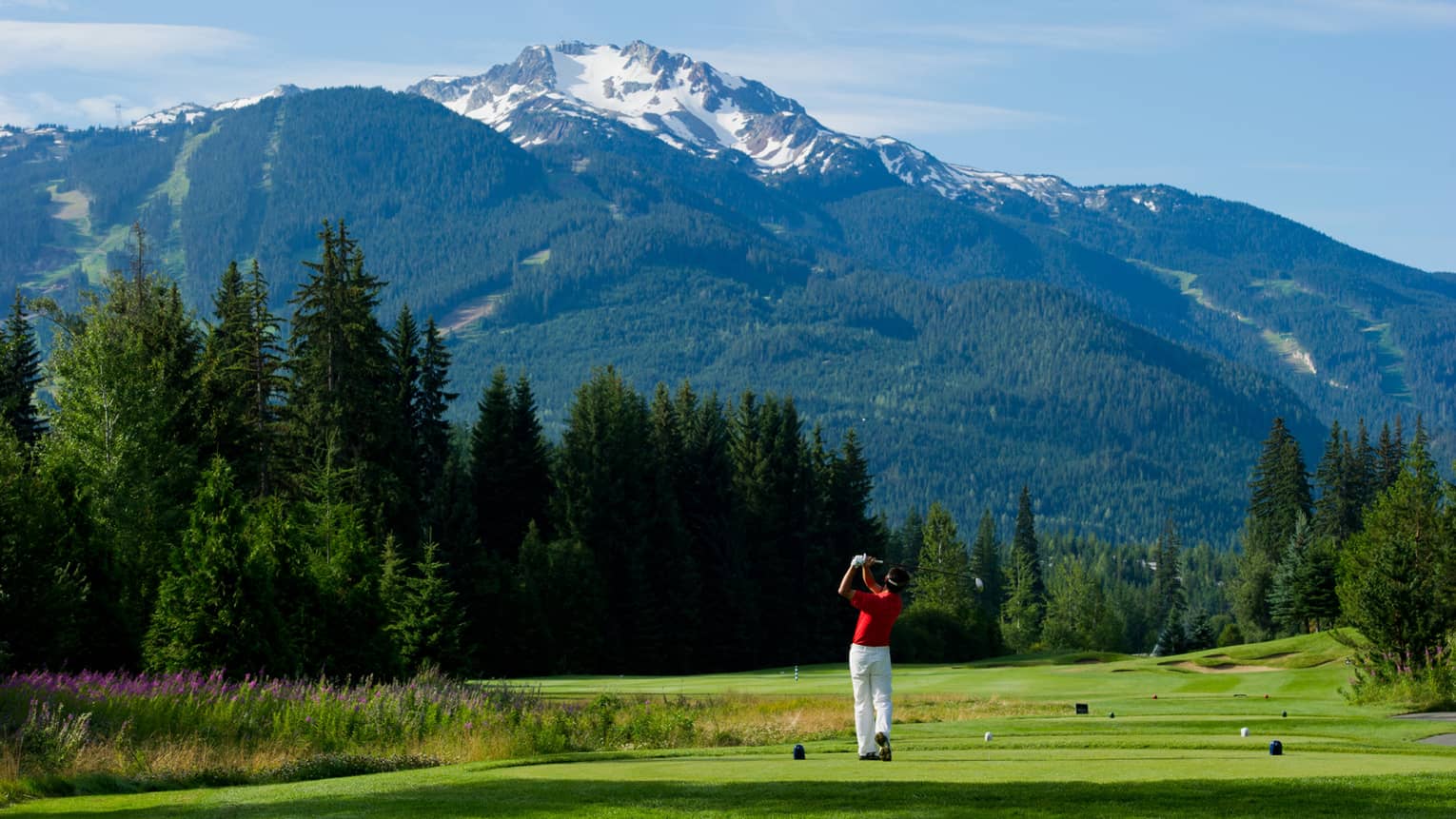 Man wearing red golf shirt swings club on green golf course surrounded by snow-capped mountains