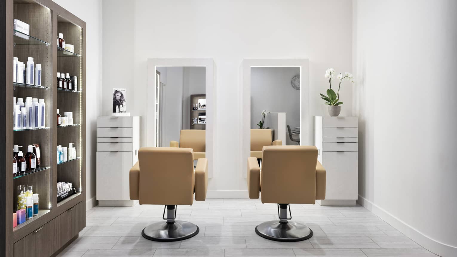 A hair salon with two light brown leather chairs facing mirrors, wall of products