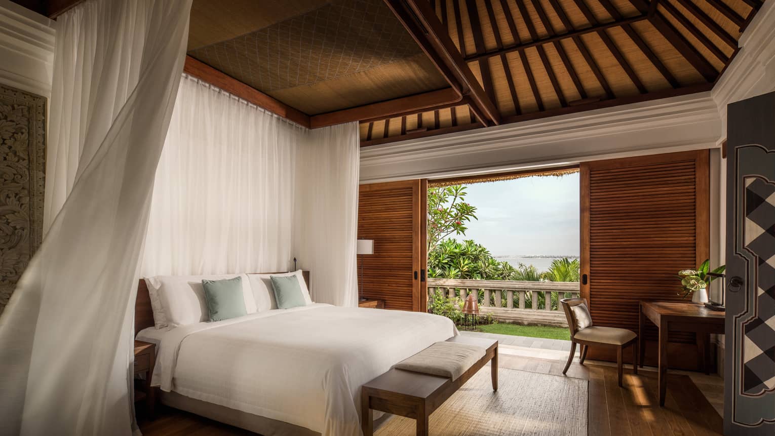Interior of resort villa bedroom with white bed and high ceilings, looking out to view of leafy green trees and ocean