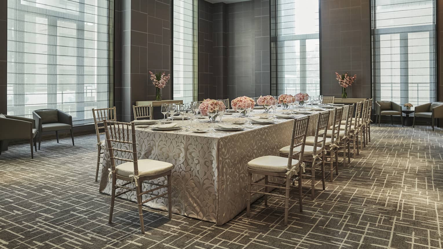 ,A long table with table cloth and flower centerpieces in a room with large windows.