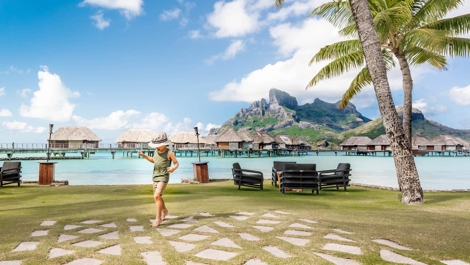 A child tiptoes on paving stones by a clear lagoon. Grass-roofed suites stand in the water in front of island mountains.