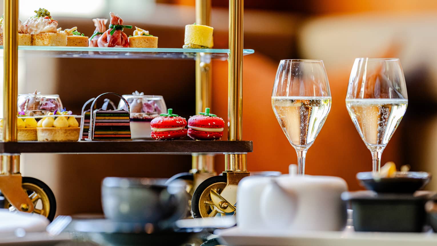 A tiered serving tray on small wheels holds colourful small bites and is sitting on a table next two two stemmed glasses holding a light-hued liquid, a teapot, and a teacup and saucer.