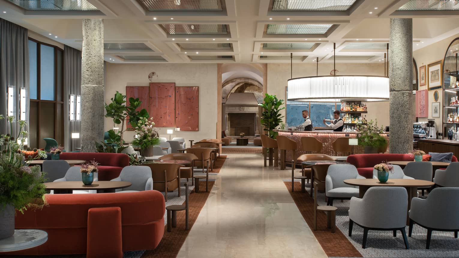 A spacious, light-filled dining area with tables, upholstered seating, potted greenery, a bar, art on the walls, and decor in beige, deep red and grey