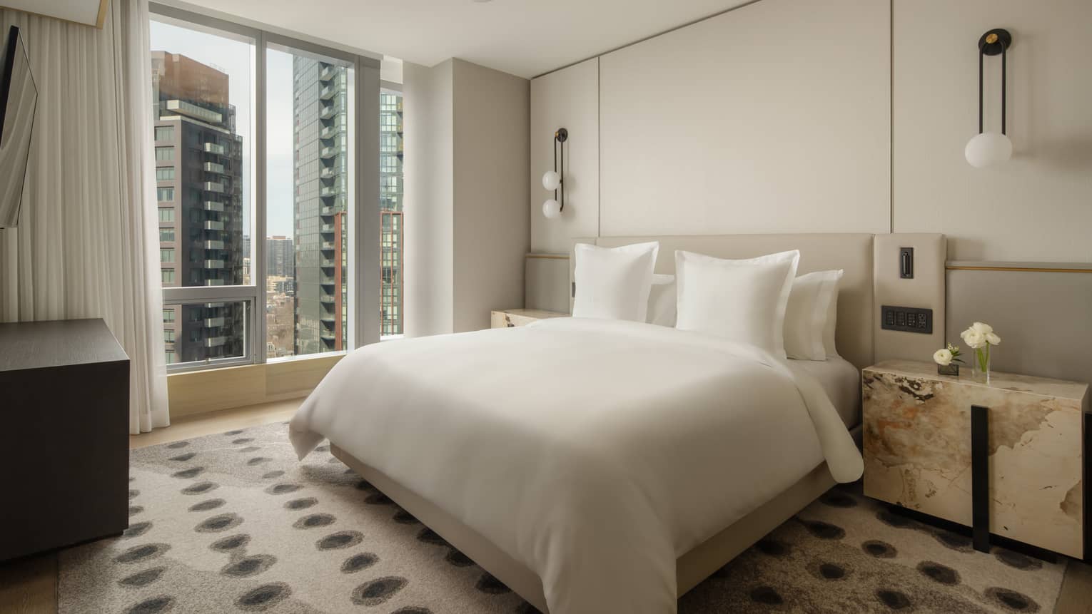 Hotel suite bedroom with a king bed, bedside lamps, nightstands and large windows overlooking city buildings