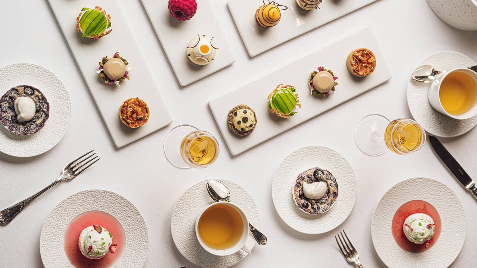 Four white rectangular platters each holding four colourful pastries each are set on a white table along with two cups of tea and four small round plates of dessert