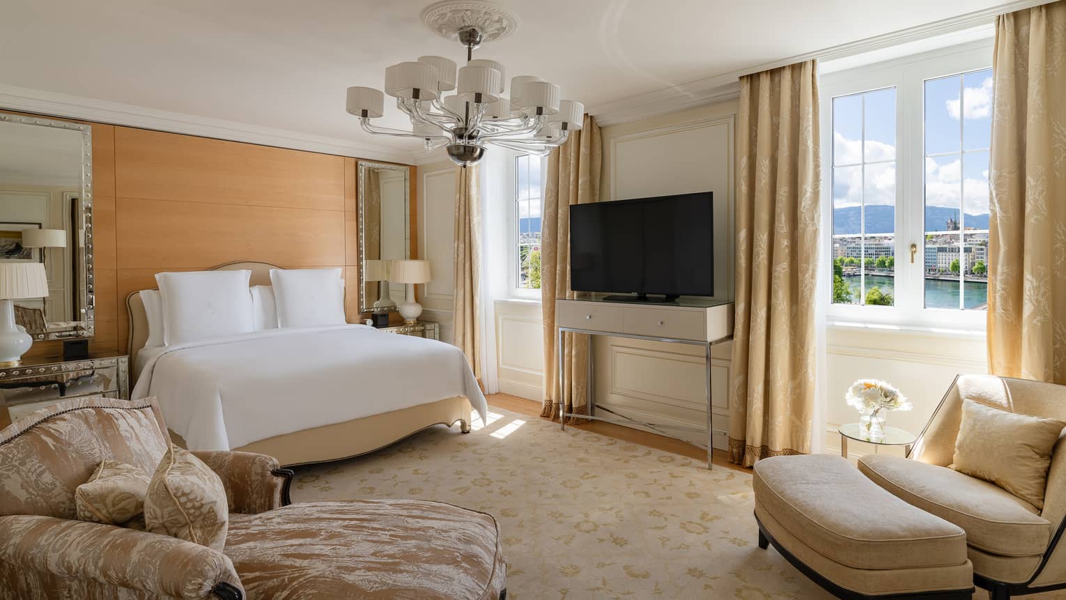 Luxury suite bedroom with a king bed, mirrored nightstands, beige décor, armchairs, ottoman, TV and windows offering a scenic view of the water and mountains