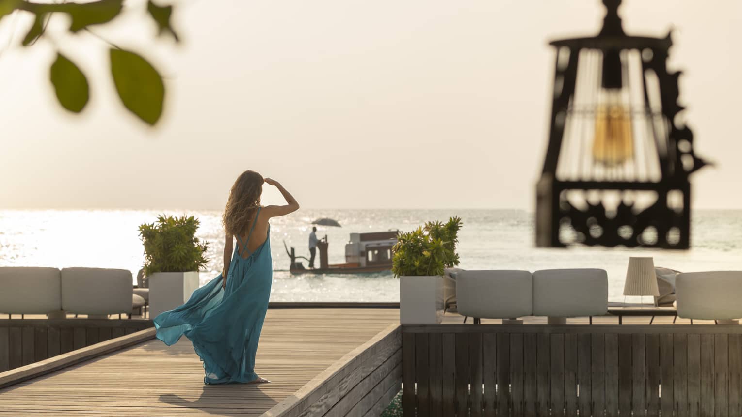 Person in flowing blue dress walks along wooden dock toward seating area at sunset
