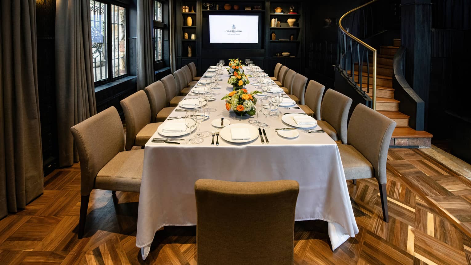 A gently lit, spacious event room with a long table set for a meal. The room has arched windows on one wall and a curved staircase on the other. There is also decorative wood flooring, wall sconce lighting and a large flat-screen TV on the end wall.