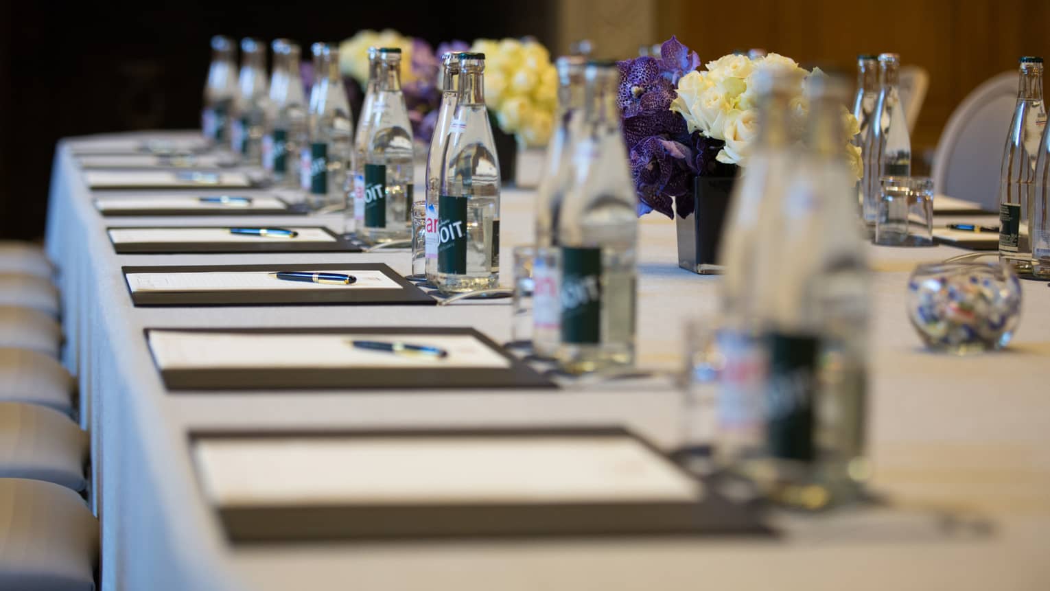 Row of meeting agendas, glass bottles, small vases with flowers on boardroom table