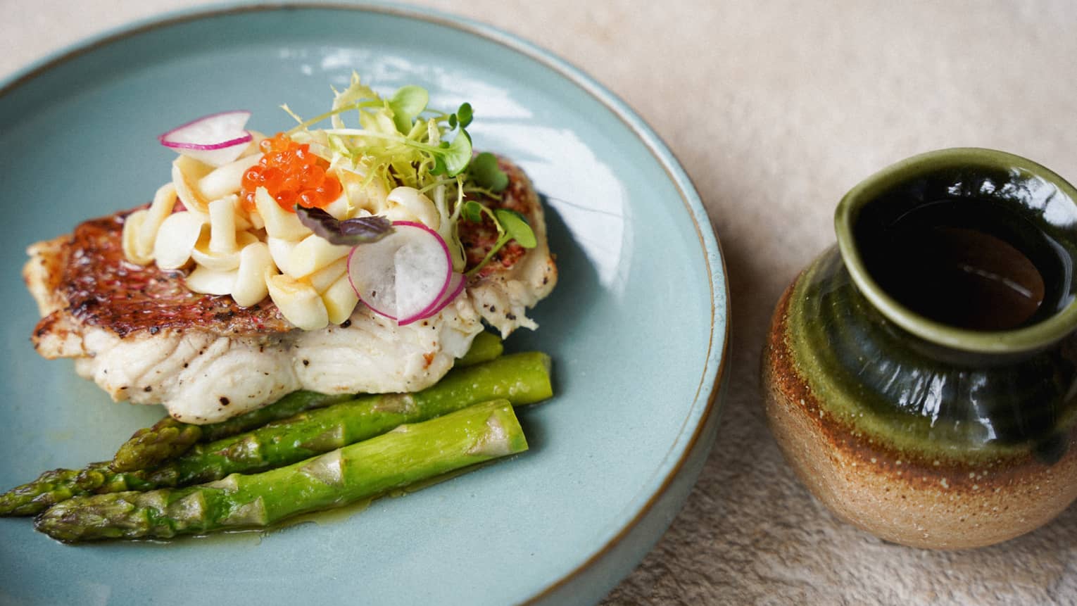 A pale blue plate holds a piece of white-fleshed fish placed next to asparagus spears and topped with garnishes. A green and tan glazed ceramic vessel sits next to the plate.