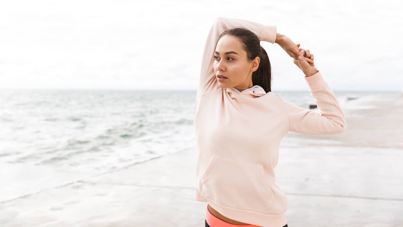 A woman in a pink hoodie stretching on a beach.