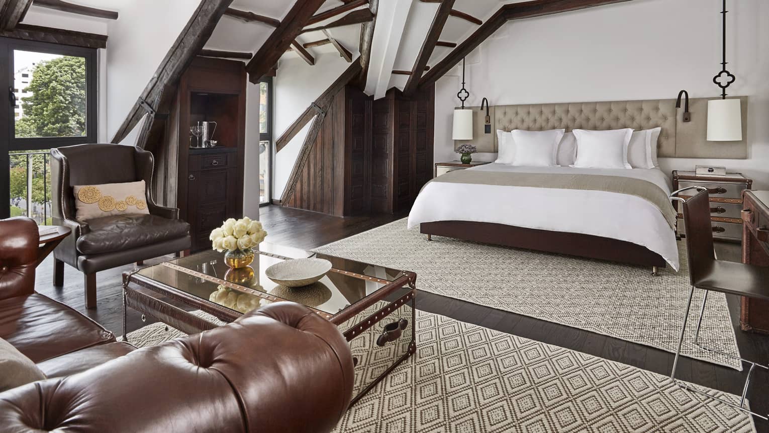 Sloped beam loft ceiling over hotel bed with padded headboard, brown leather armchair, table with flowers