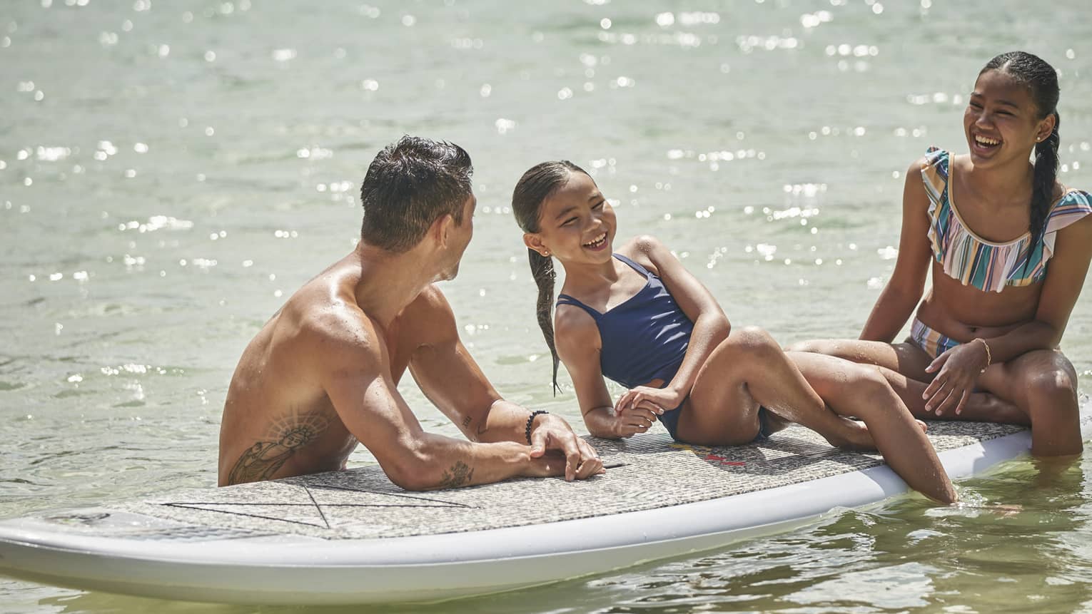 Two children sit on a stand-up paddleboard smiling as an instructor looks toward them holding the board still in the water