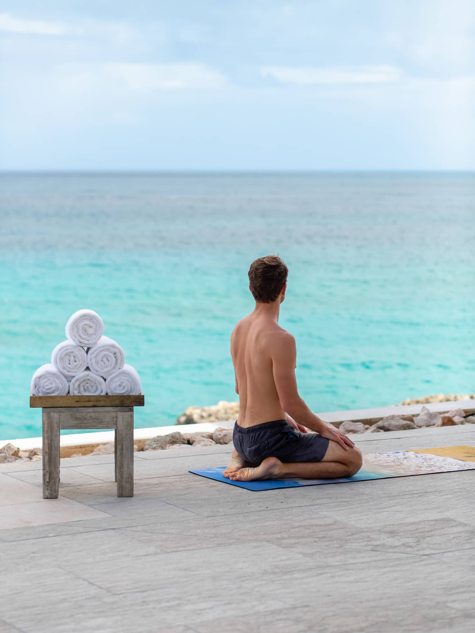 A man sitting on his legs with the ocean in front of him and towels on a bench behind him.