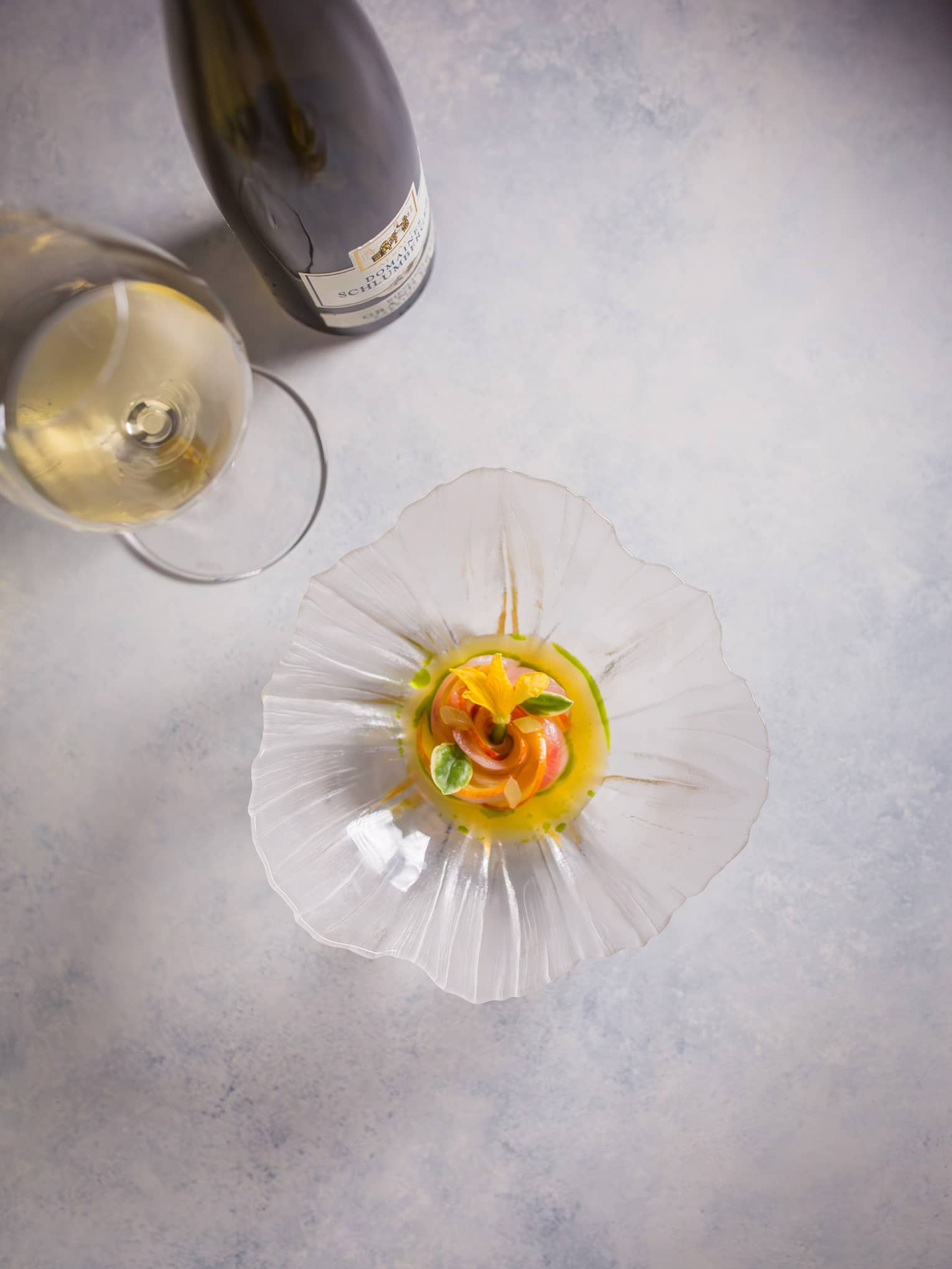 Overhead view of a colourful appetizer garnished with florals in a glass, flower-shaped dish beside a glass of white wine.