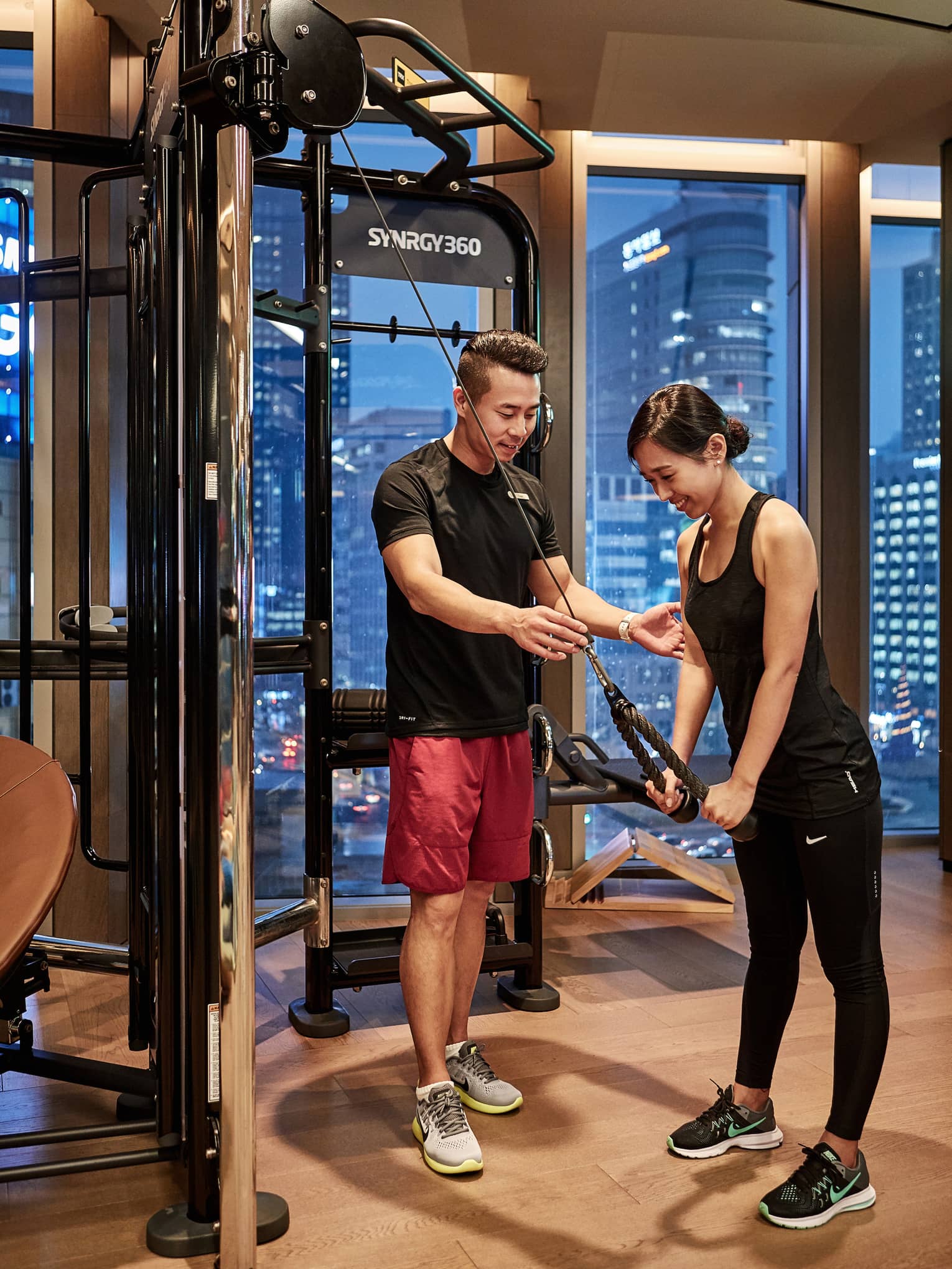 With skyscrapers out the window beyond, a trainer guides a guest as she performs a triceps pushdown exercise on a gym machine.