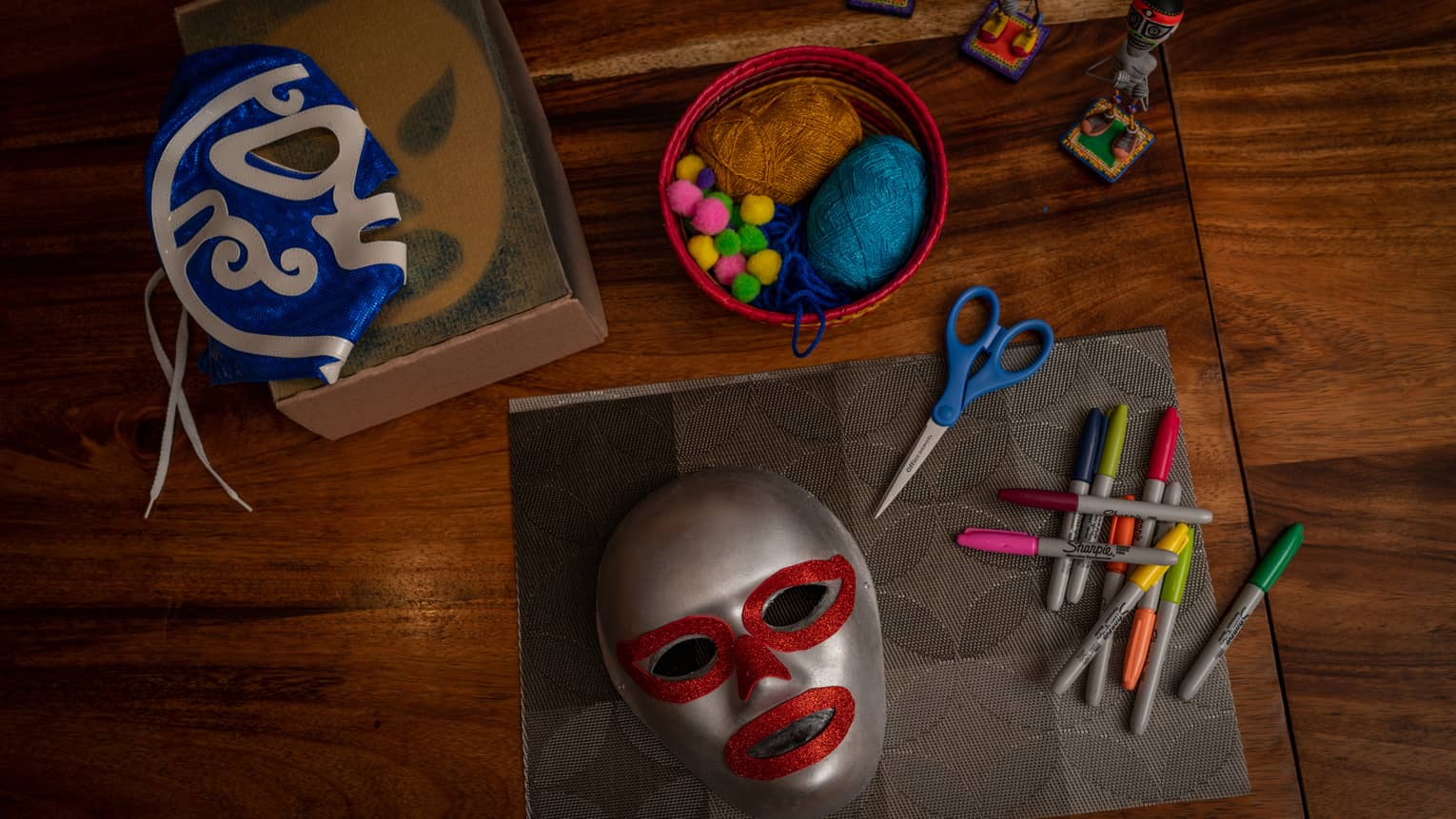 Atop a wooden surface, small figurines, masks and mask-making supplies: scissors, colourful markers, pompoms and yarn.