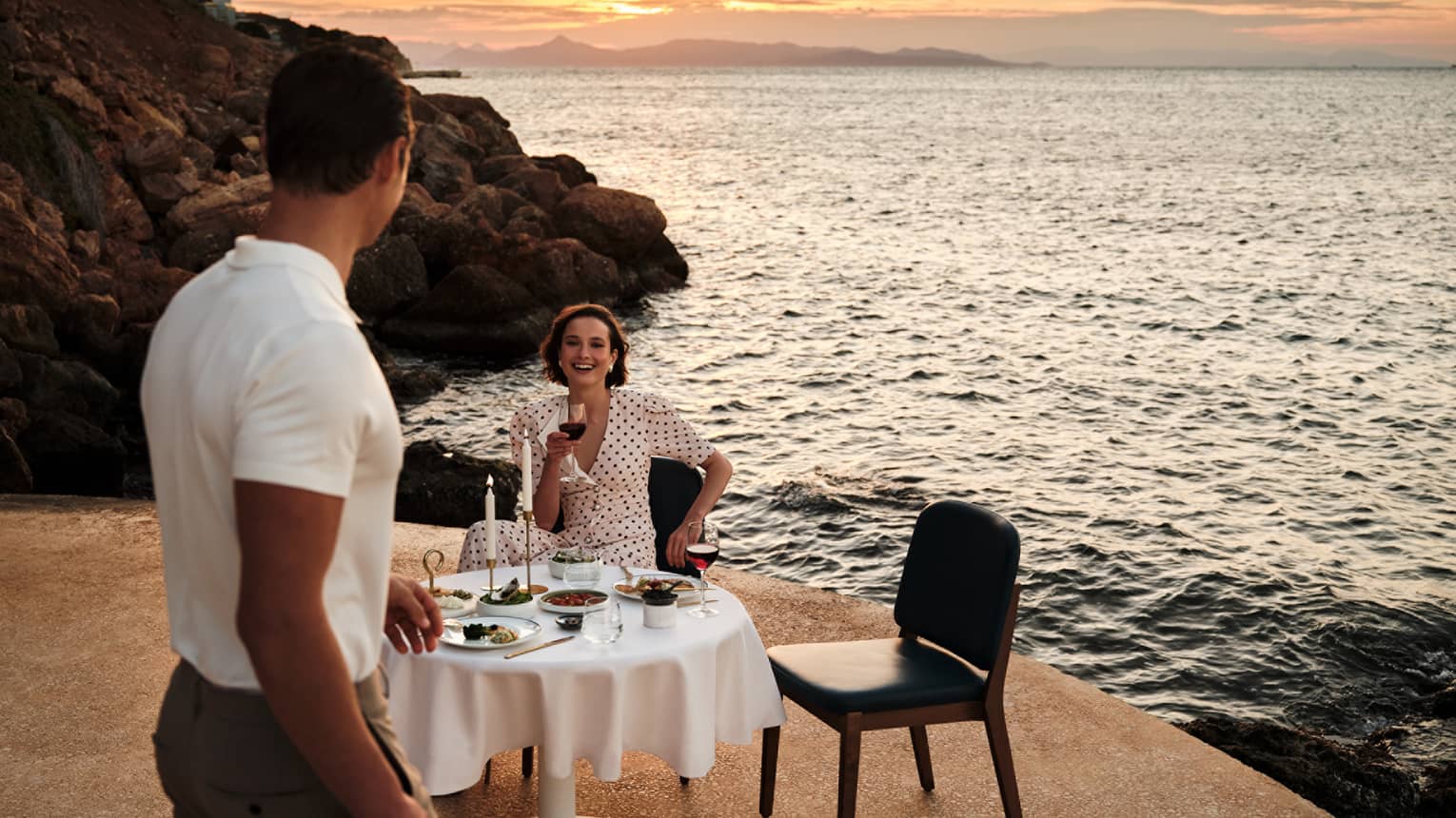 A person sits at an elegant dining table in an outdoor setting, while another person walks towards the table. A rocky shoreline, the setting sun and the ocean are in the background.