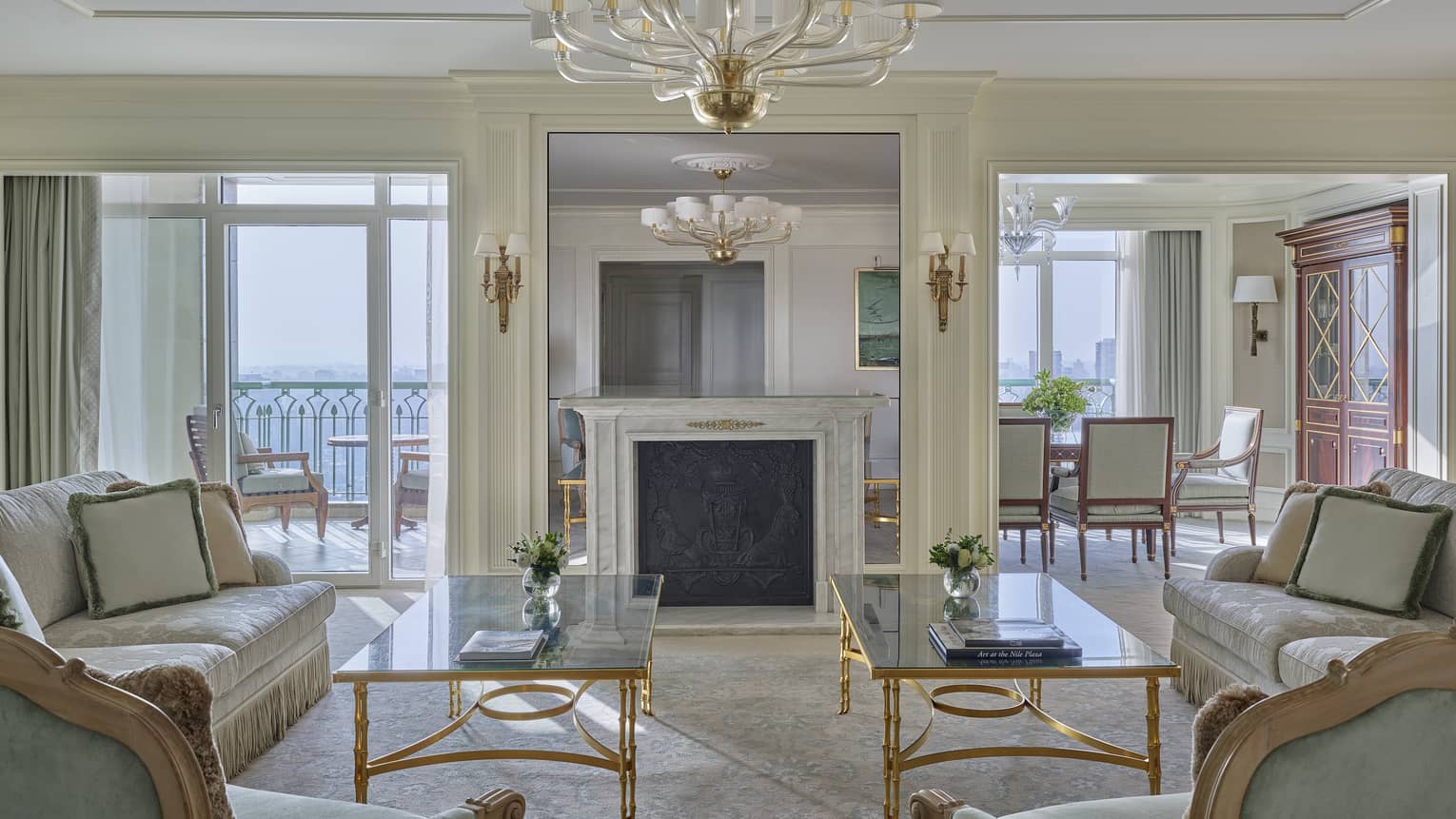 Elegant hotel suite living room featuring two glass coffee tables, two sofas, two arm chairs, chandelier, fireplace and walk-out balcony