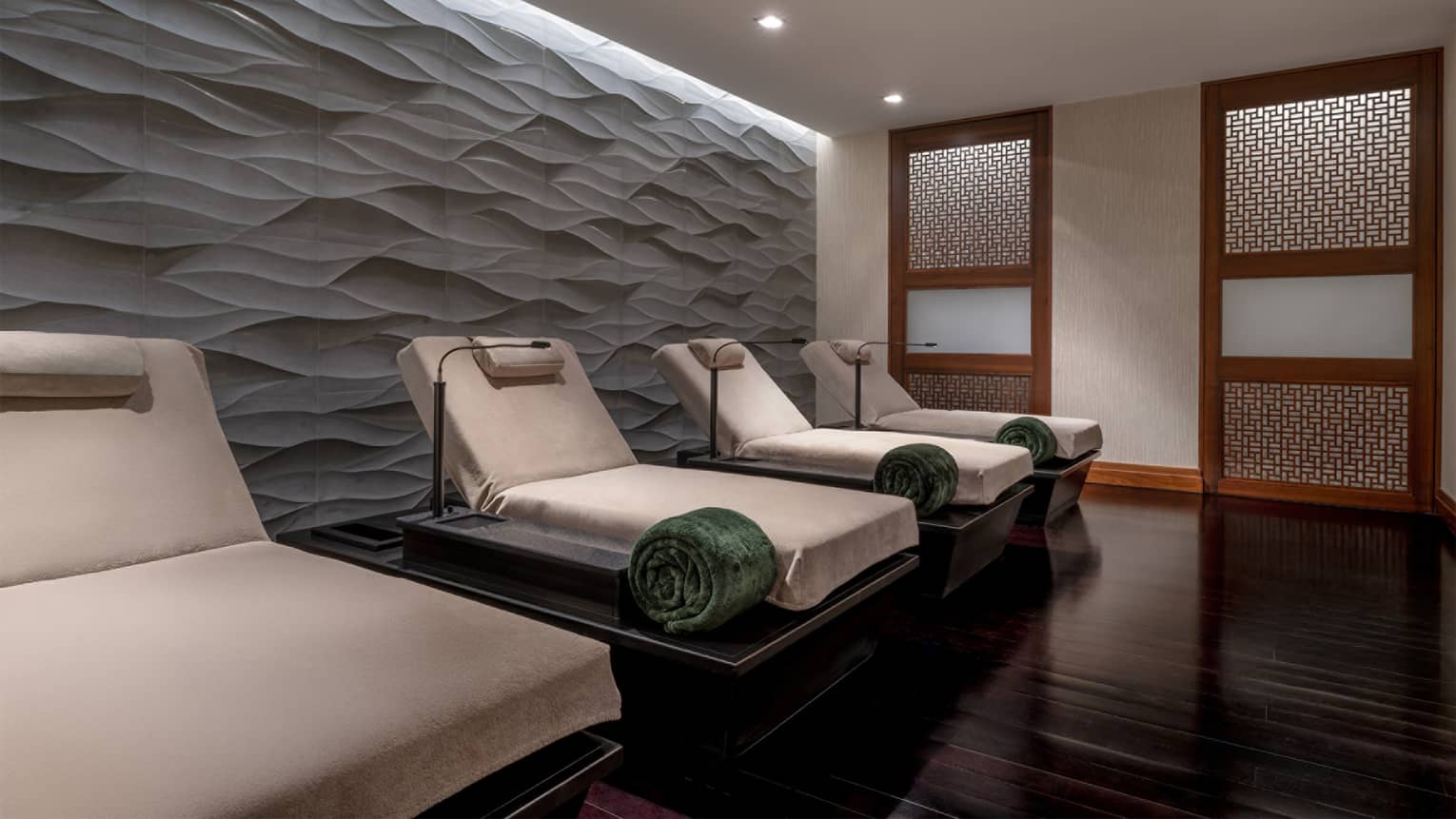 Spa relaxtaion room with a row of four upholstered lounge chairs in natural linen, each with a green rolled up blanket, a head pillow and a small lamp, all set in front of a textured wall