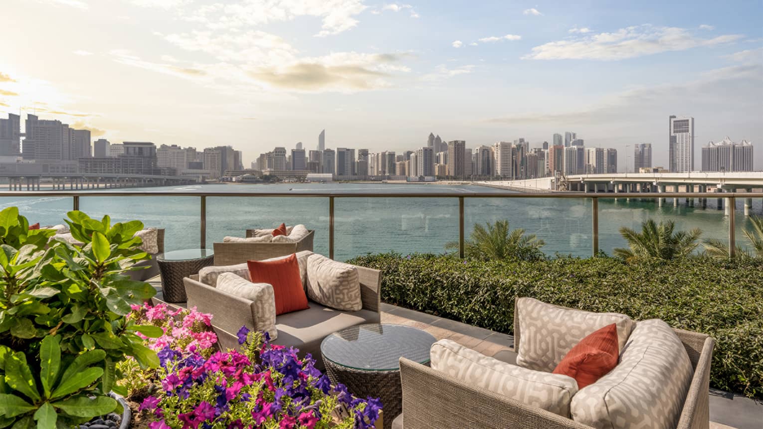 Open-air terrace with two light-grey conversation sets surrounded by greenery and flowers and overlooking the water with a city skyline in the background