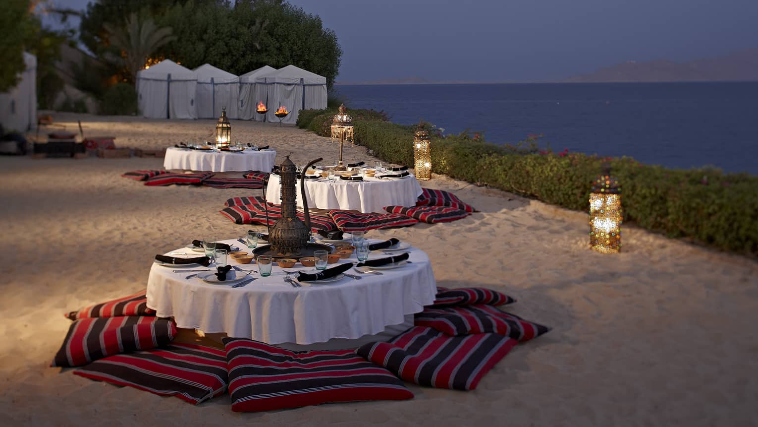 Round dining tables with cushions on sand beach with lanterns at night