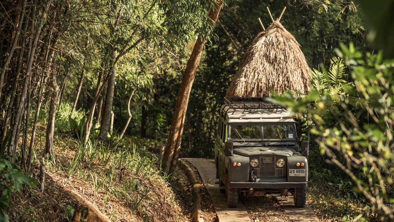 Jeep driving down path in forest past thatched-roof hut