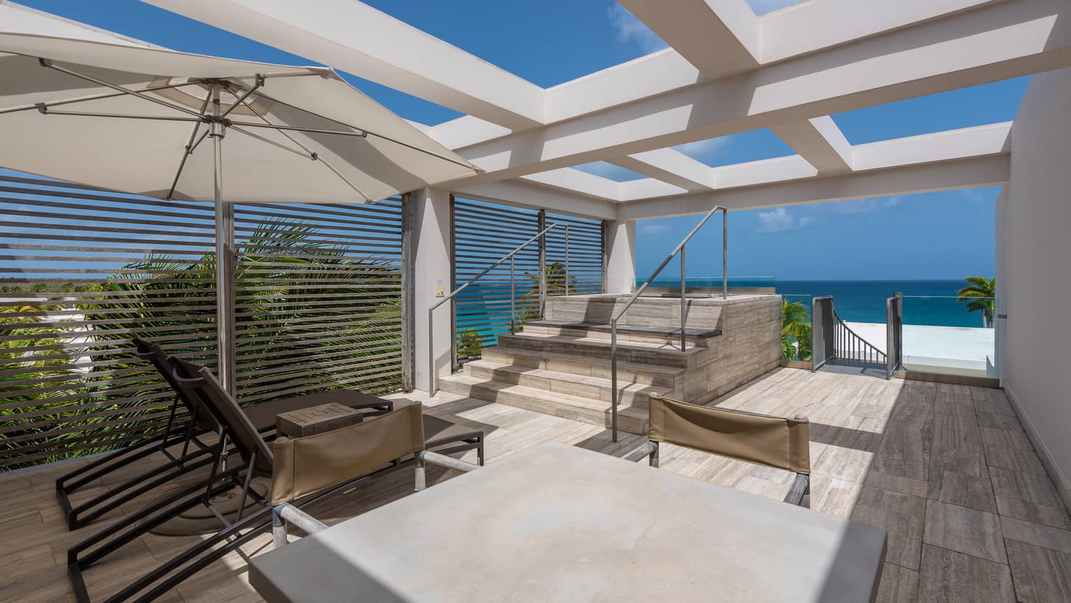 Resort terrace with lounge chairs, a sunshade, stone steps, and a view of the ocean and palm trees under a clear blue sky