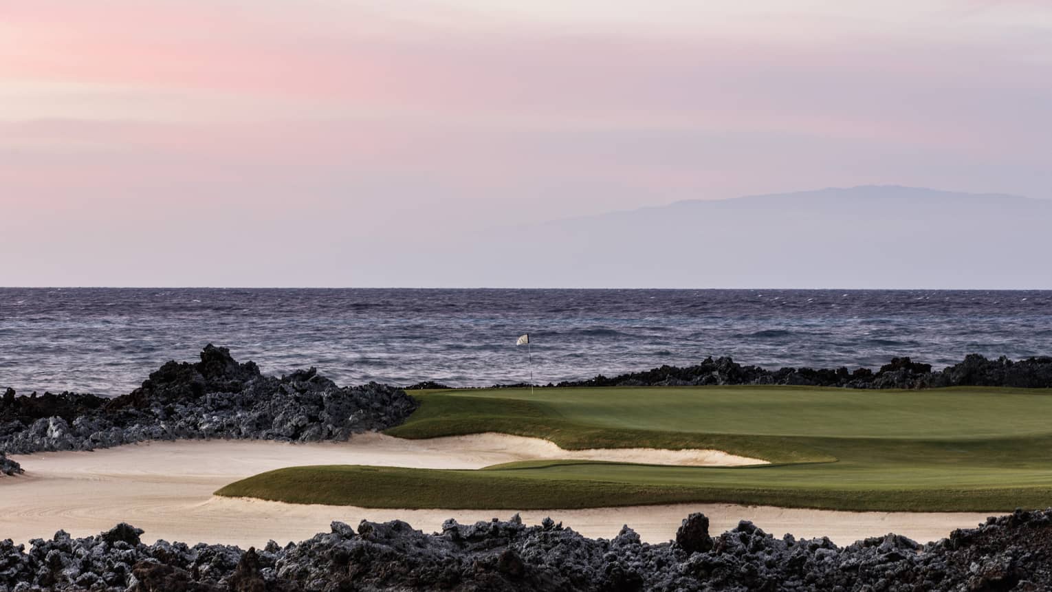 On an oceanside golf course beneath a rose-coloured sky, a single flagstick stands amid black lava rocks and white sand. 