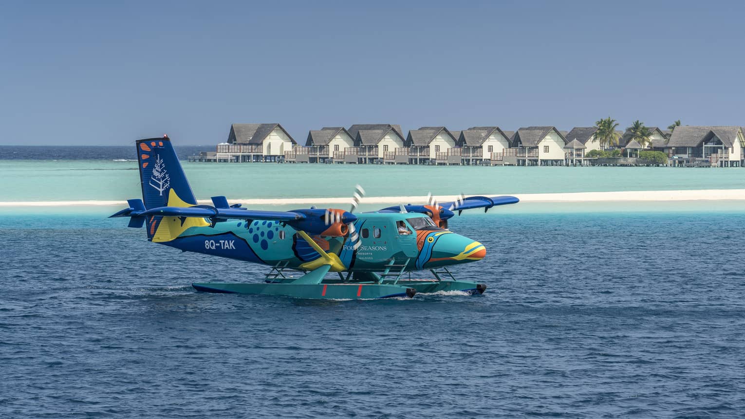 The Four Seasons sea plane landing on calm water in front of beach houses and palm trees