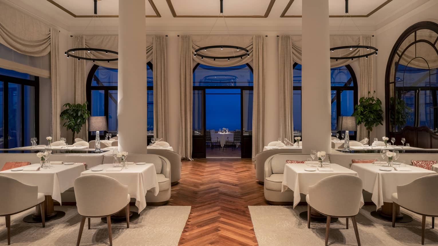 A restaurant with columns, large windows and white cloth tables.