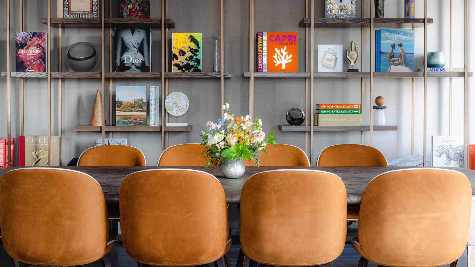 Rectangular table with floral centerpiece and eight suede chairs, copper bookshelf with books and trinkets