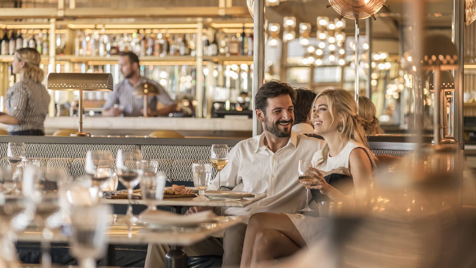 Smiling couple drink wine in a booth of a luxurious restaurant decorated in an elevated neutral and bronze colour palette.