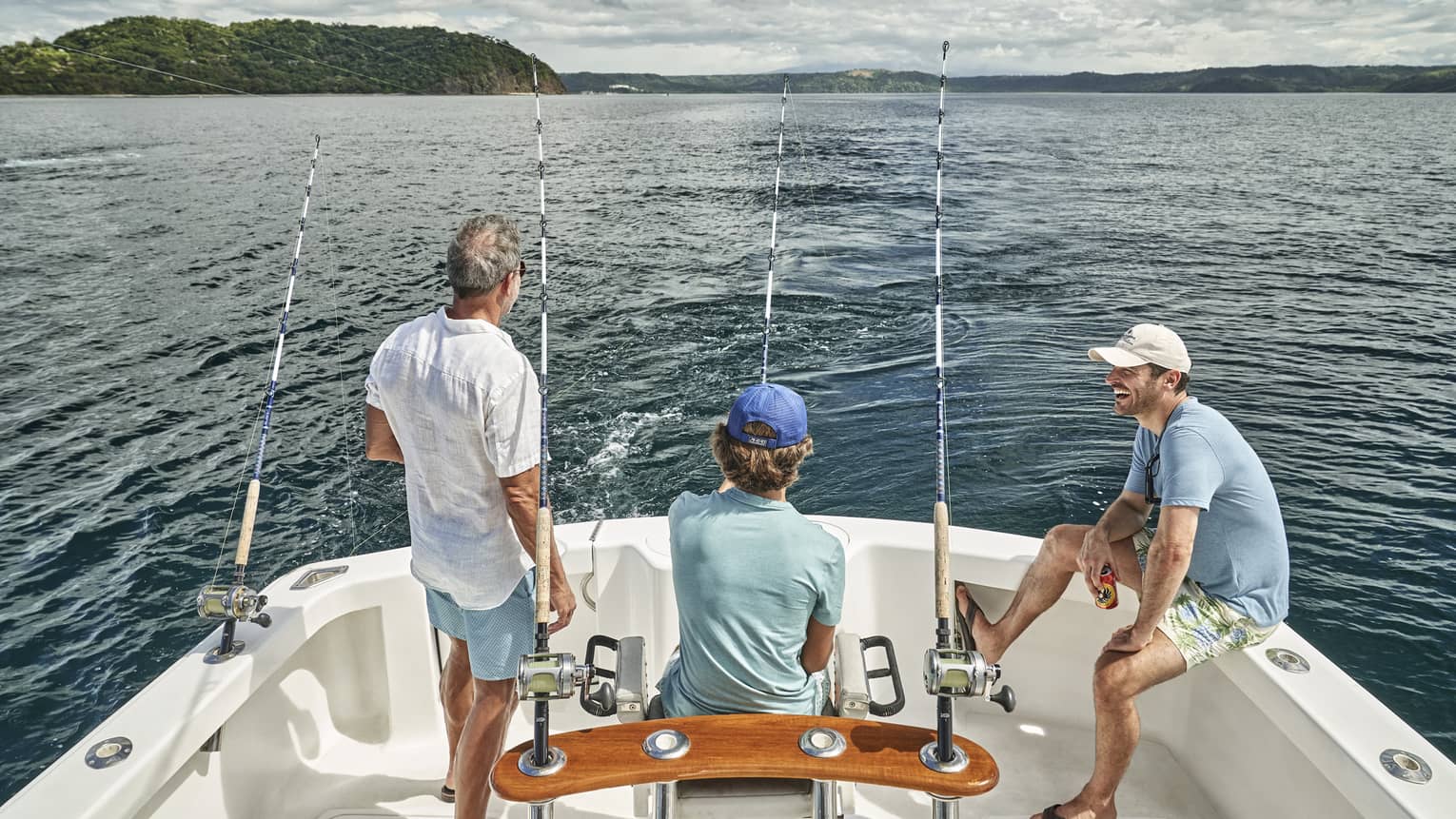 Three men of varying ages sit looking out at the water from the back of a boat outfitted with fishing rods