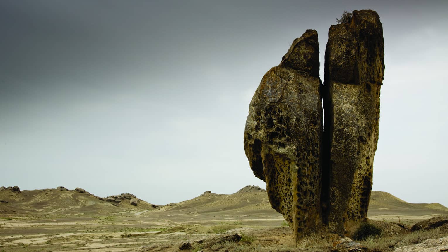 A rough stone formation proudly juts upwards toward a moody grey sky against a background of arid, rolling brown hills.