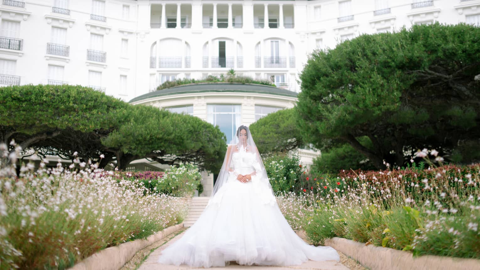 Bride wearing wedding gown and long sheer veil over face stands in garden before Four Seasons Hotel Grand-Hotel du Cap-Ferrat