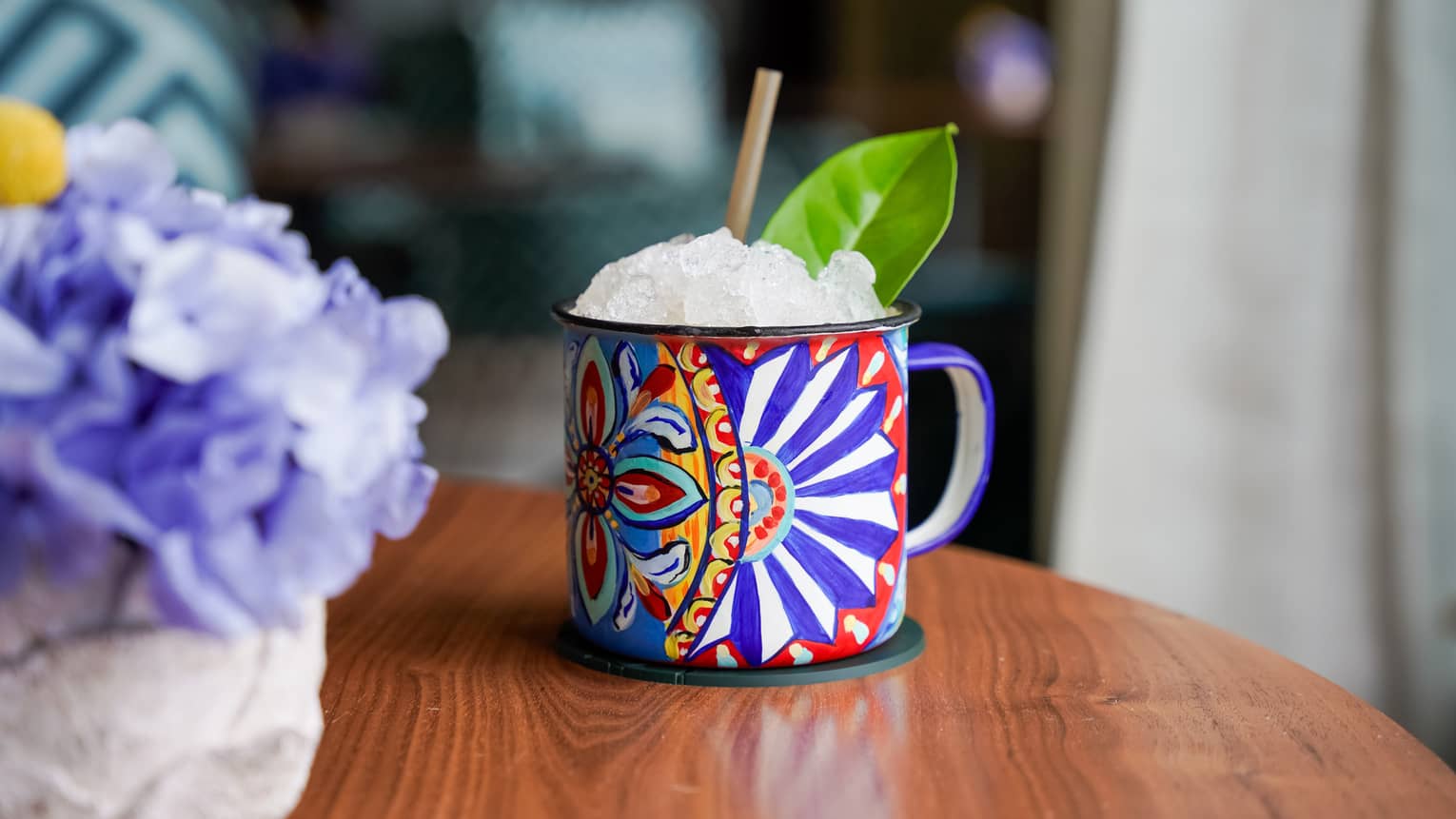 ,A colourful patterned mug filled with ice and garnished with a green leaf sits on the edge of a round wooden table