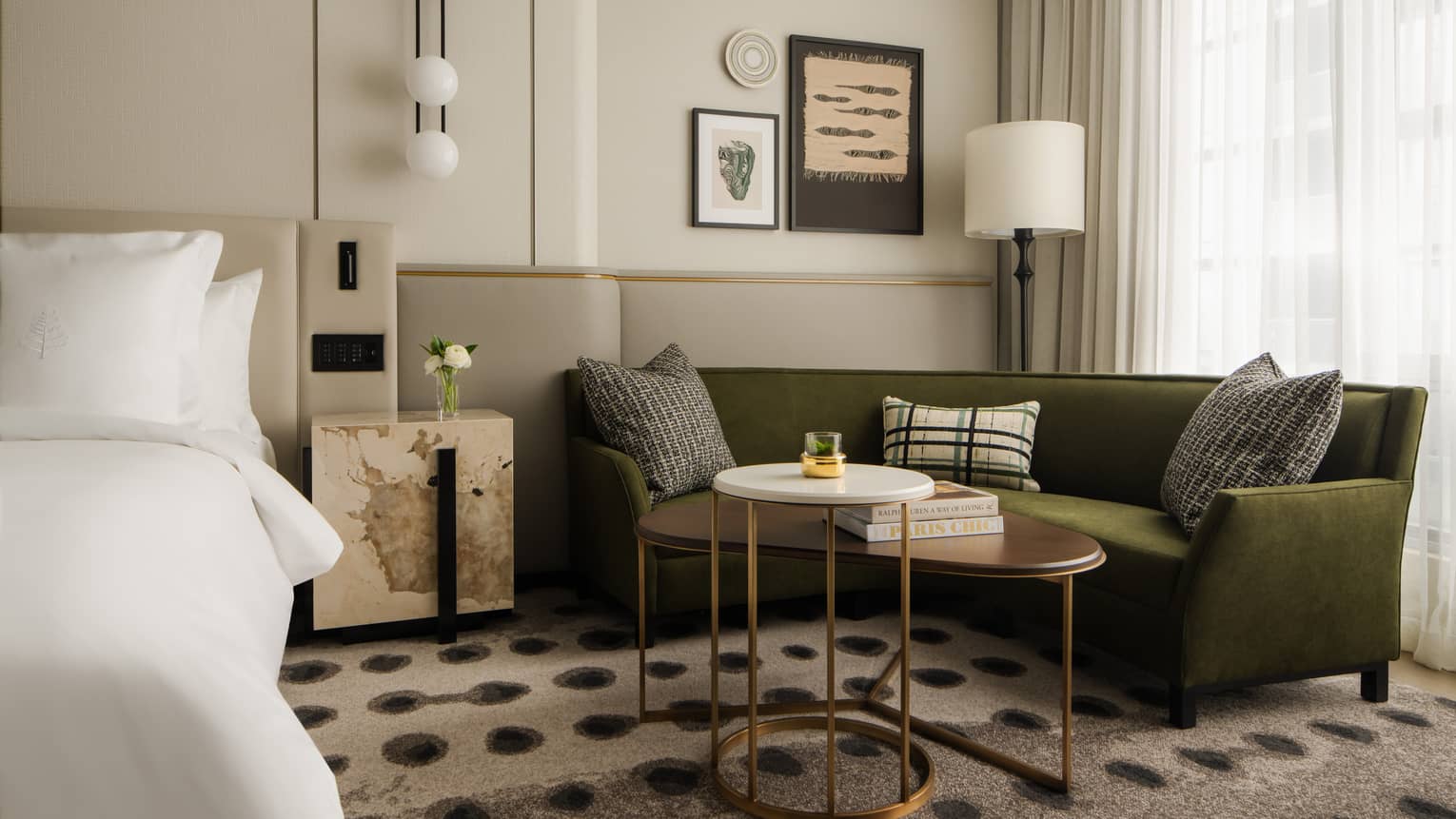 Hotel room lounge area with a green sofa, decorative cushions, modern artwork, adjacent bed and large windows 