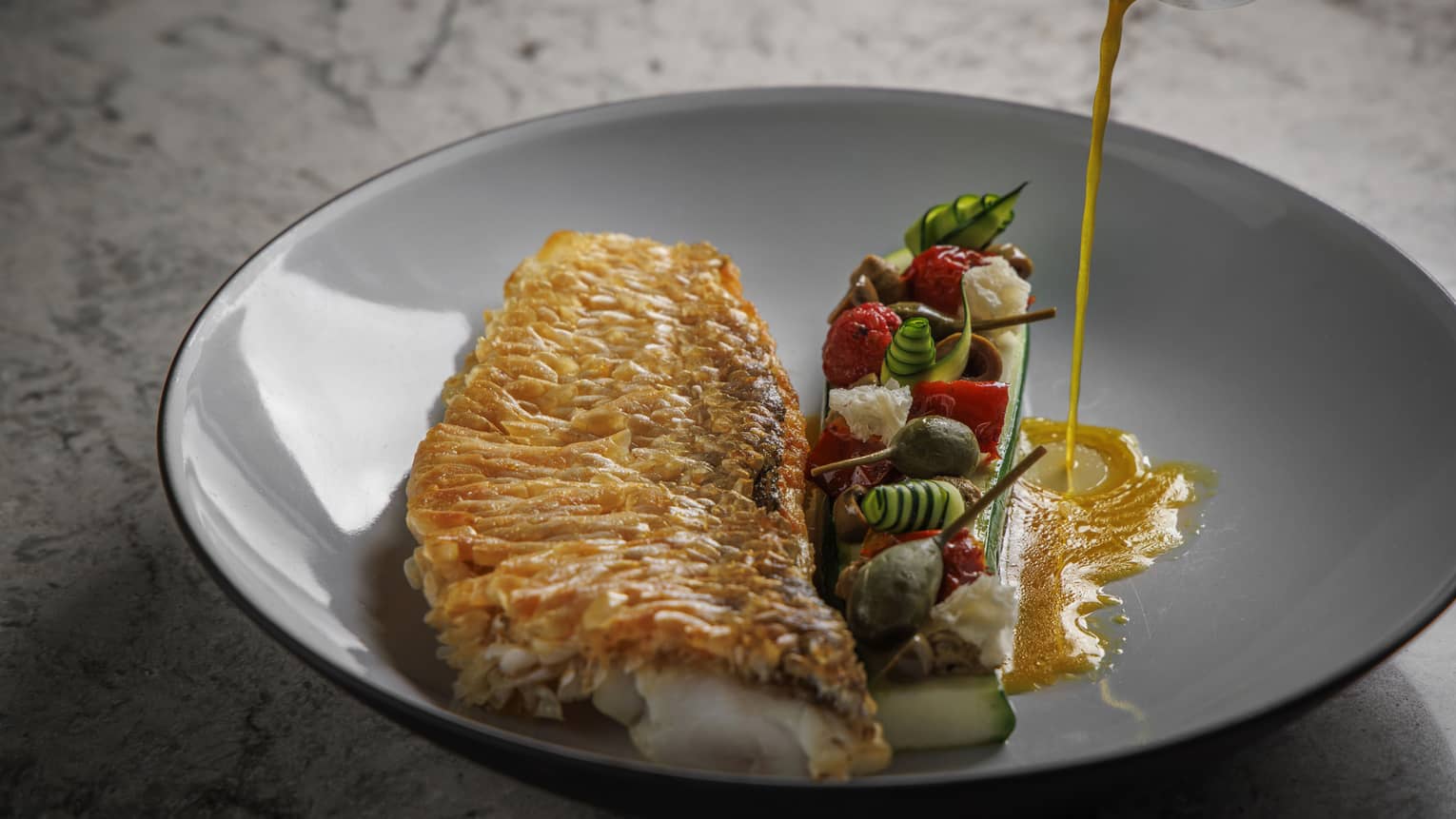 Fish filet with assorted vegetables in shallow grey bowl, sauce being poured into it