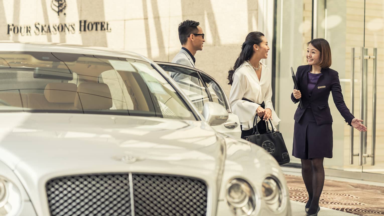 A person in a dark suit greets a couple near a luxury white car at the hotel entrance. The couple appears enthusiastic and engaged in conversation.