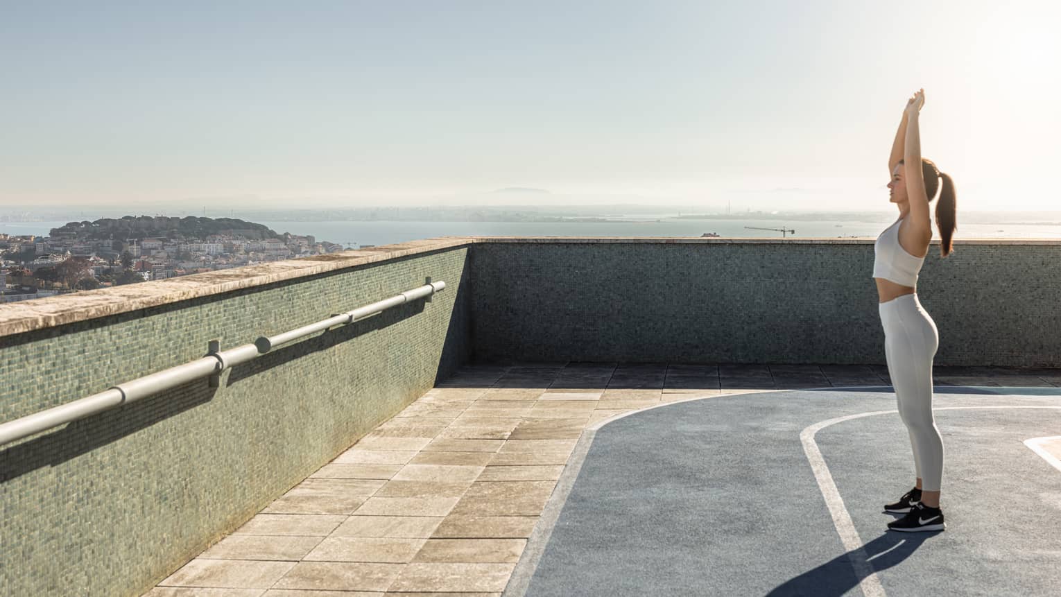 A person stretches with arms overhead while standing on a rooftop running track on a sunny day, with the horizon between water and sky visible in the background