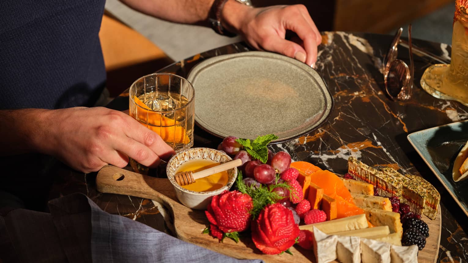 A charcuterie board with fruits and cheeses on a table beside a drinking glass