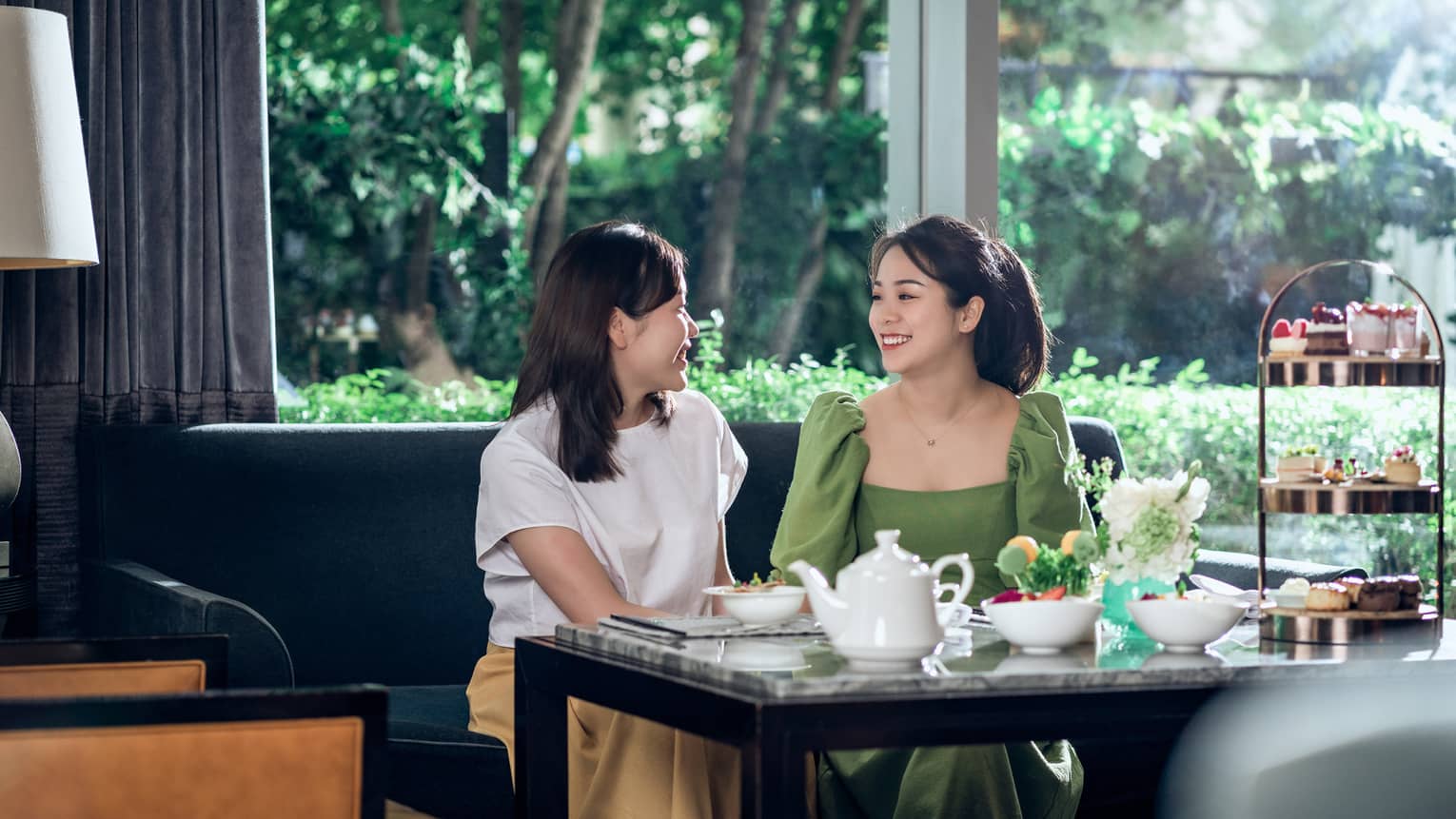 Two women sitting and smiling over afternoon tea in front of large window