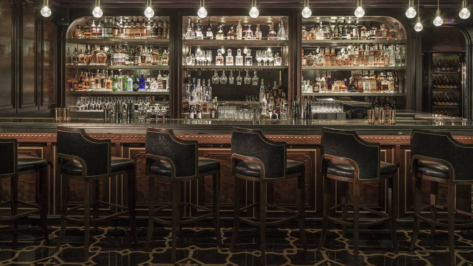 Dimly lit bar counter with black leather bar stools in front and mirrored shelving holding various bottles of spirits behind the bar