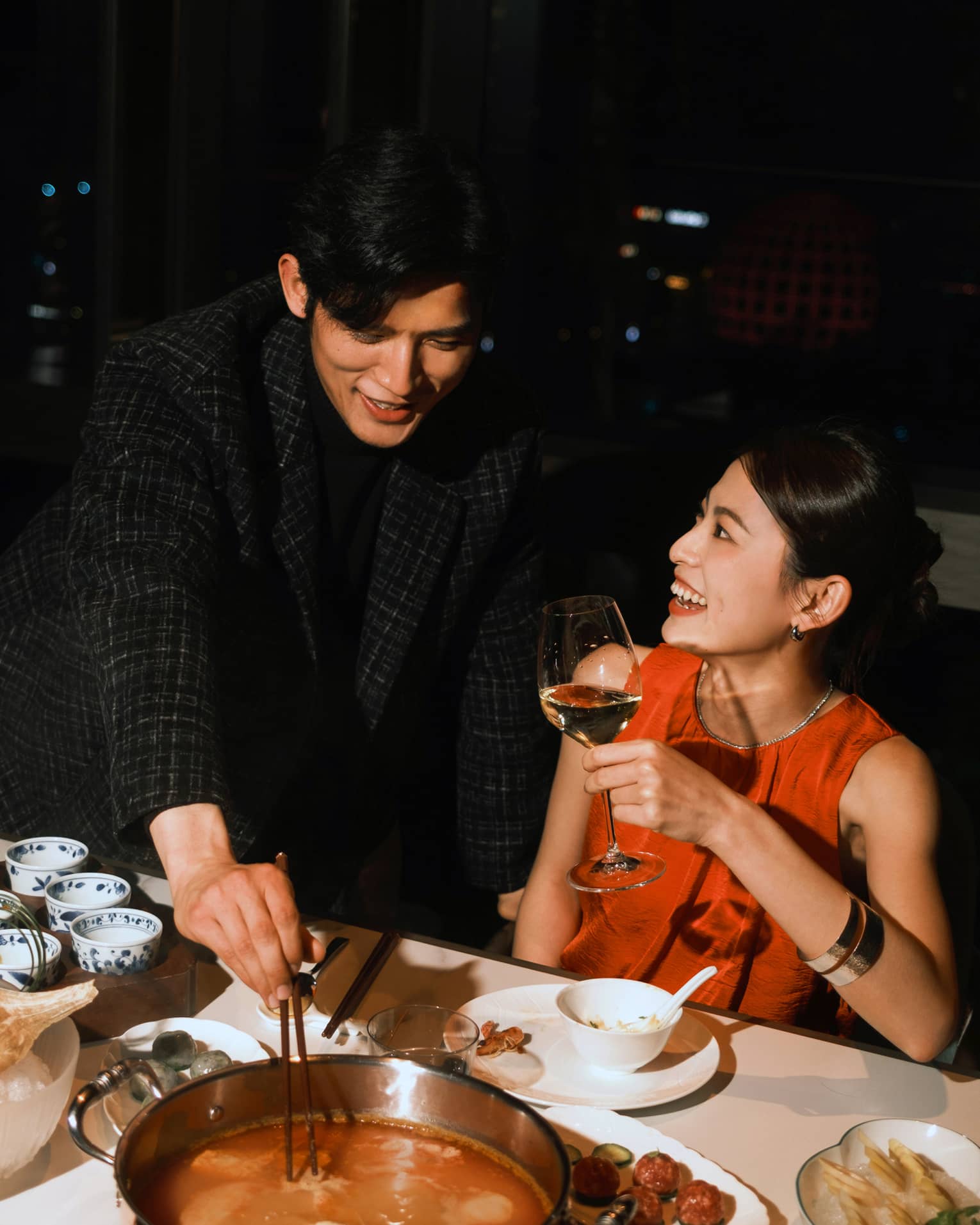 Guest reaches into a hot pot with chopsticks while another smiles holding a glass of white wine. Behind them is a night sky.