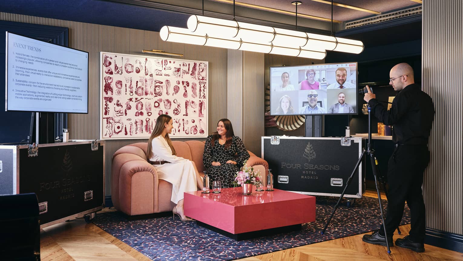 Two people sit on a couch in a well-lit meeting space with wood flooring and an area rug. Another person stands in front of them with a camera on a tripod. Art hangs on the wall behind the couch, and the couch is flanked by two flat-screen monitors. A coffee table is in front of the couch.