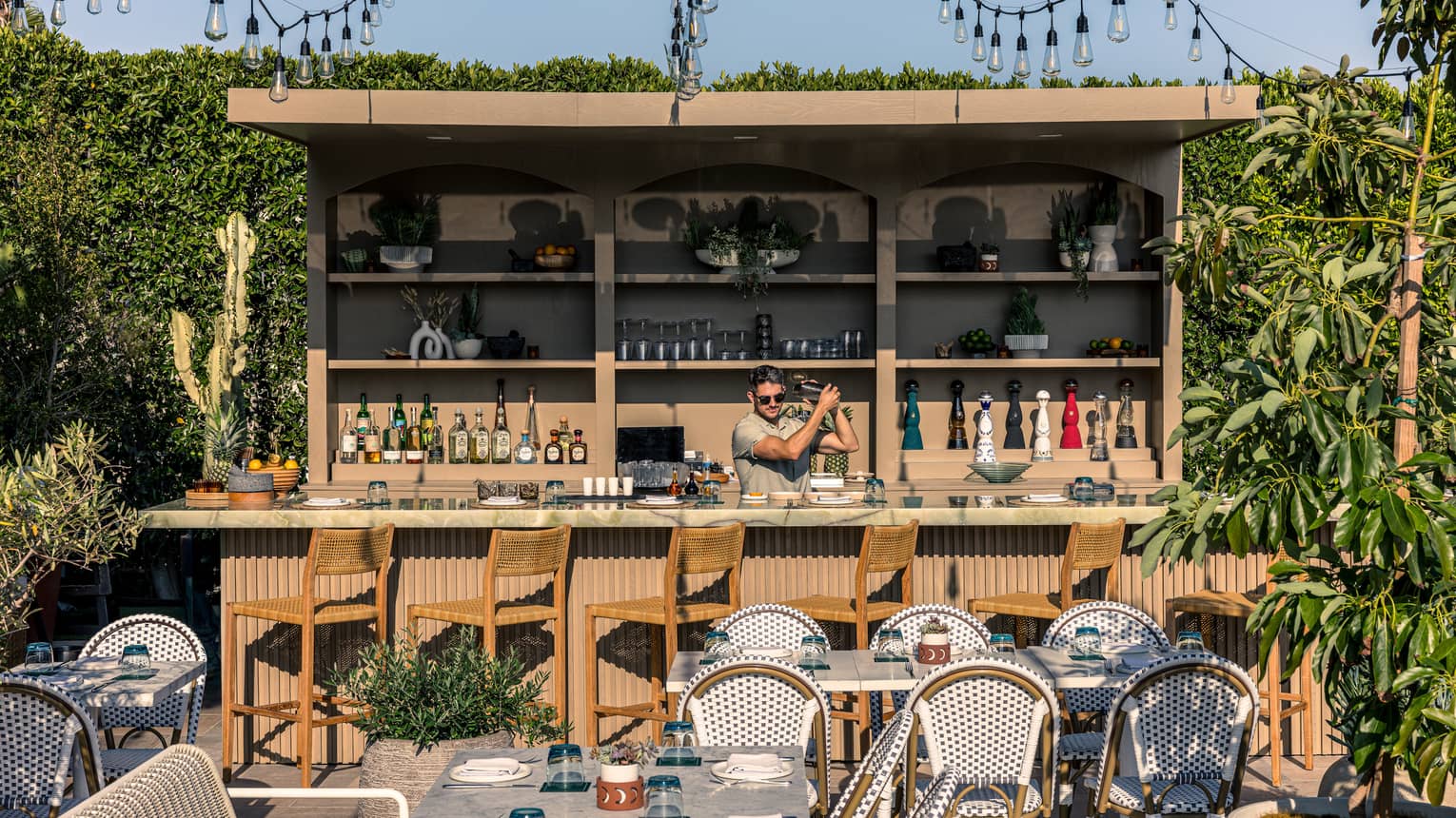 Outdoor restaurant patio with bartender standing behind a wood bar counter with shelving behind them and sting lights overhead