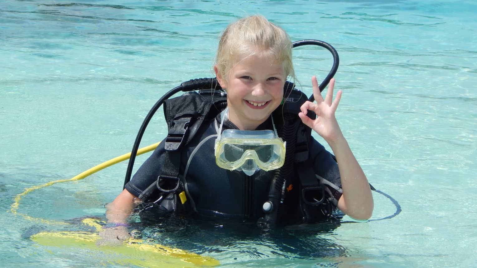 Young girl wearing scuba diving gear in water smiles, gives hand signal