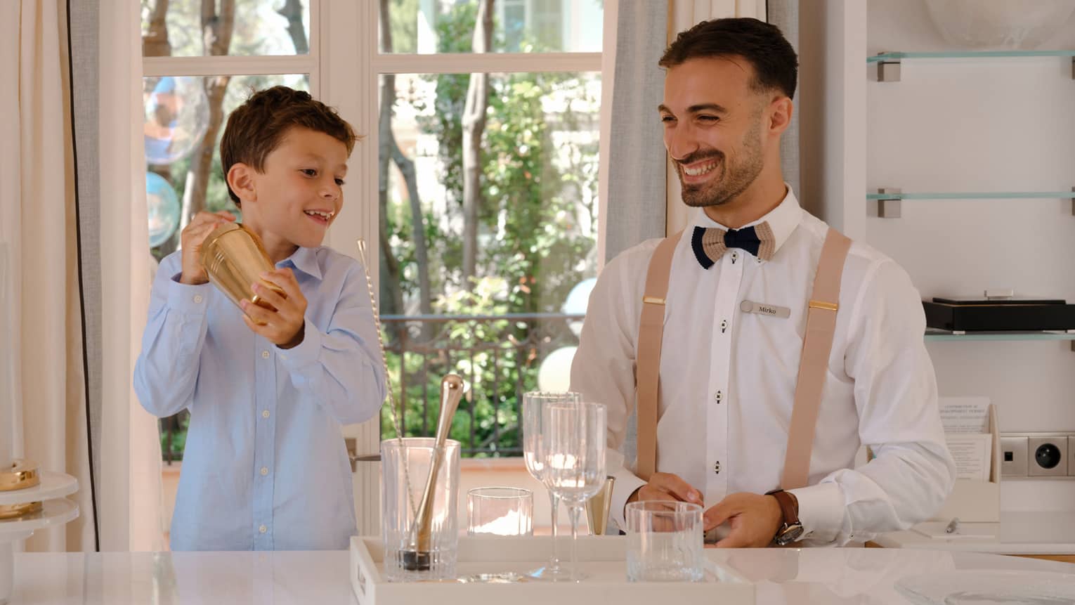 Child holds gold mocktail shaker as smiling bartender looks on